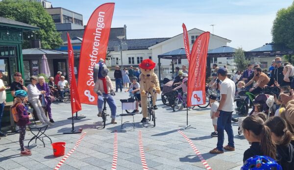 Syklistforeningens maskot rulle og Brannvesenets maskot Bjørnis i en balanse-duell på sykkel med mange tilskuere i alle aldre