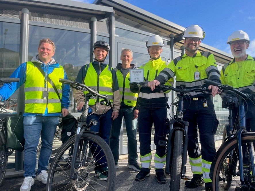6 menn med gule vester og hjelmer som poserer smilende med en sykkelvennlig arbeidsplass-plakett